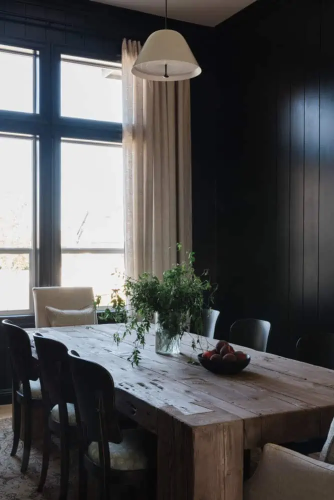 angled view of a dining room with black walls, rustic wood table, and a large window with white linen drapes