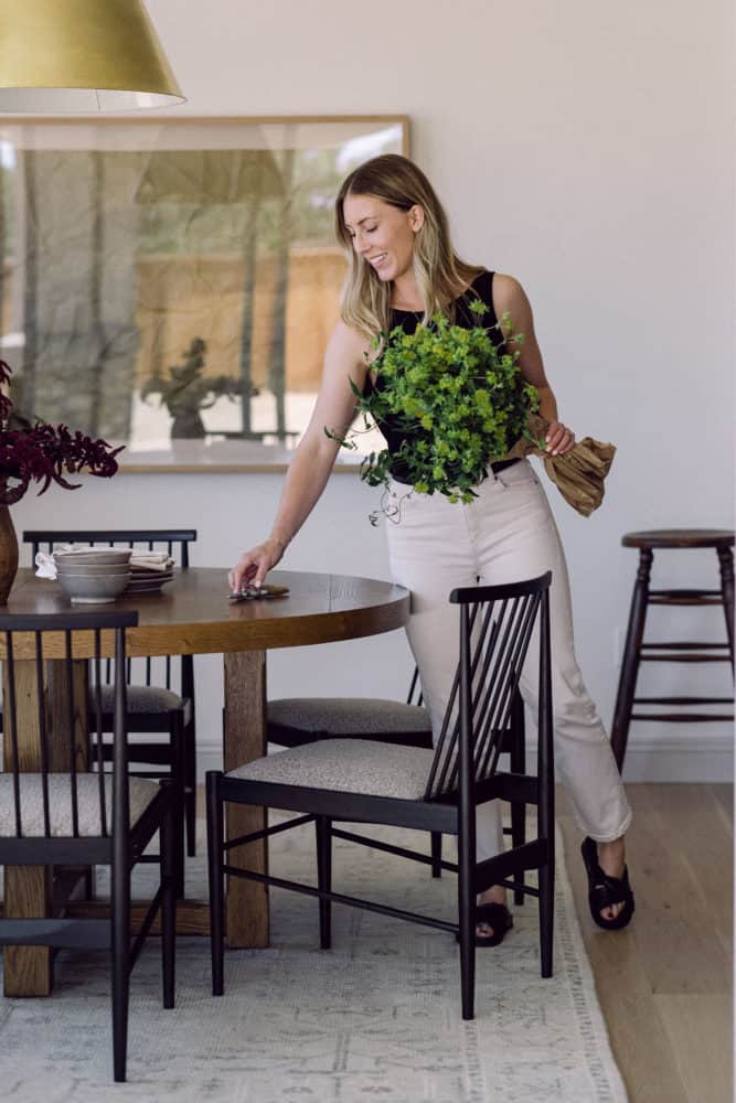woman leaning over round wooden table with plant stem in her hand with a dining chair in the foreground.