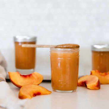 3 ball canning mason jars with peach ginger butter inside on a white countertop with fresh peach slices