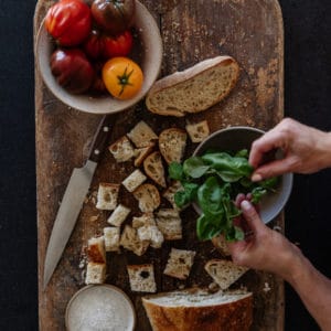 rustic cutting board on black granite with cubed sourdough, bowl of tomatoes, and hands tearing some fresh basil