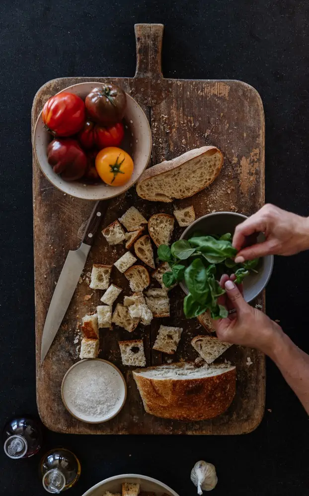 rustic cutting board on black granite with cubed sourdough, bowl of tomatoes, and hands tearing some fresh basil