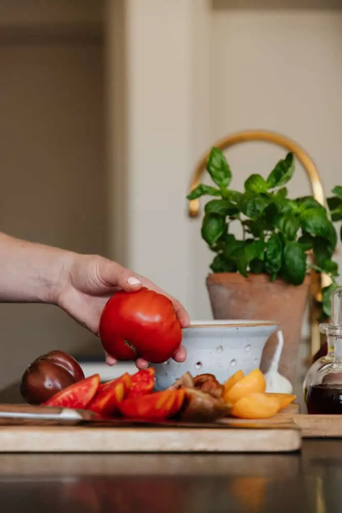 hand grabbing a fresh tomato with cut tomatoes on a cutting board with fresh herbs in a vase in the background