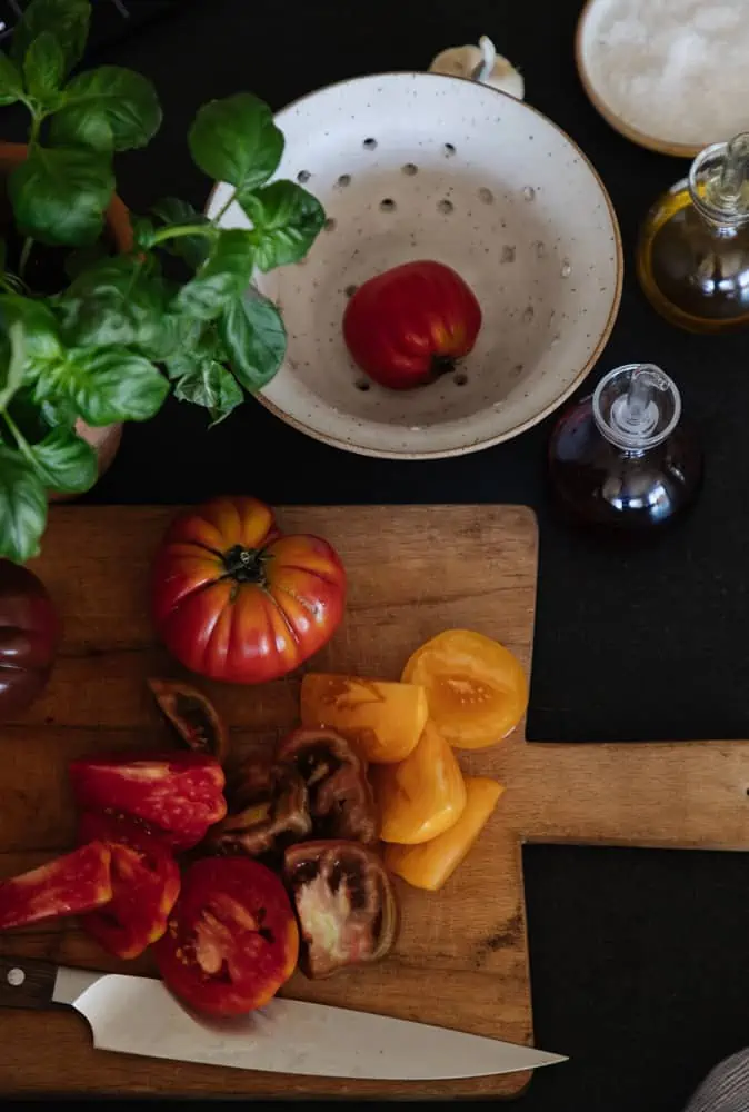 top view of heirloom tomatoes being sliced on a rustic wood cutting board with a colander and basil plant