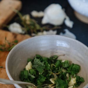 close up of fresh herbs in a bowl with sliced garlic and a fork with a cutting board and a garlic bulb in the background