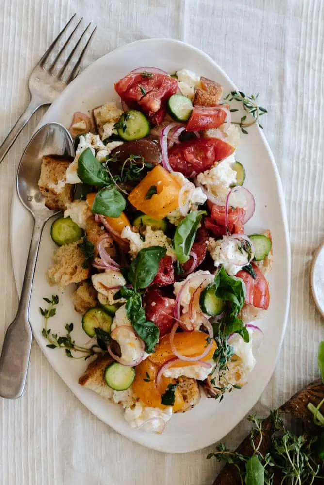 panzanella salad on a white platter on a linen tablecloth with a fork and knife leaning on the platter