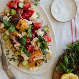 panzanella salad on a white oval platter on top on a linen tablecloth with a salt pinch pot in the top right and fresh herbs and lemon in the bottom right.