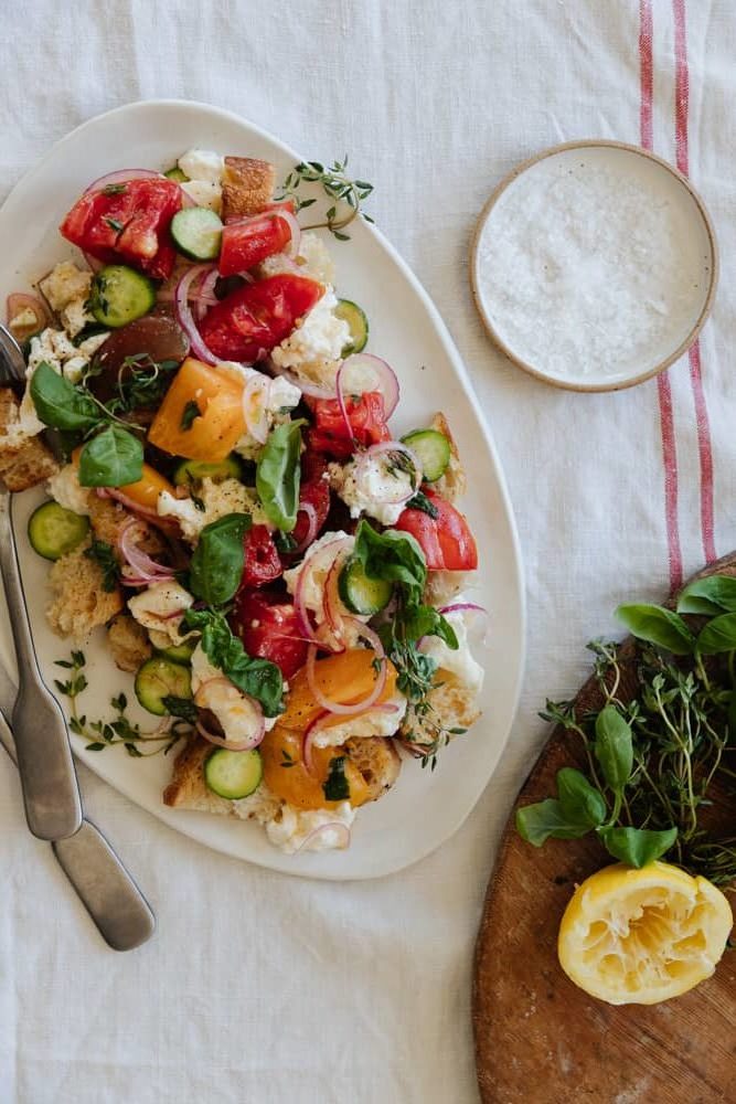 panzanella salad on a white oval platter on top on a linen tablecloth with a salt pinch pot in the top right and fresh herbs and lemon in the bottom right.