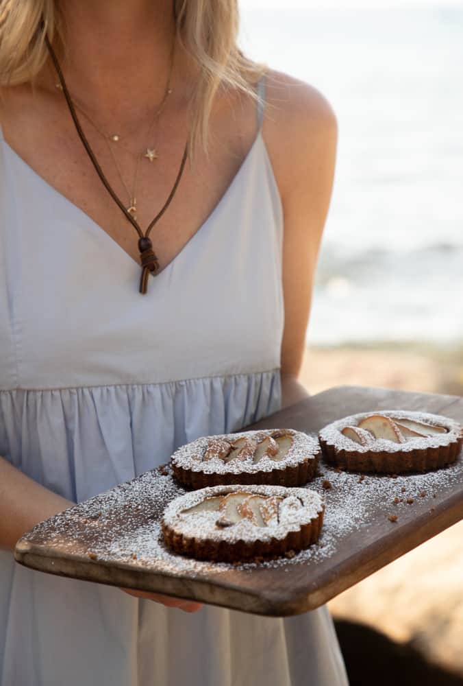 woman in a dress holding a rustic wood tray with 3 small pear tarts with powdered sugar dusted on top