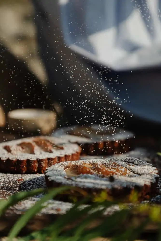 powdered sugar being dusted on top of 3 small pear tarts on top of a rustic wood cutting board