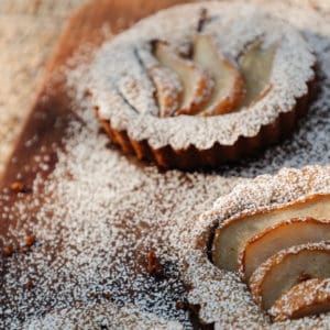 3 small pear topped tarts with powdered sugar sitting on a wood cutting board on top of a knit blanket