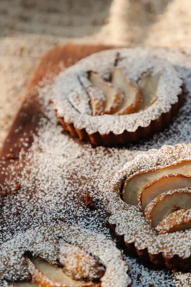 3 small pear topped tarts with powdered sugar sitting on a wood cutting board on top of a knit blanket