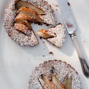 top view of 2 pear tarts with powdered sugar on a white plate with a knife to the right and one slice cut out