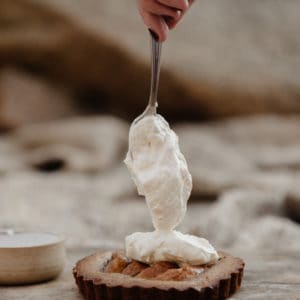 womans hand holding a spoon vertically above a pear tart with whipped cream being dolloped on top