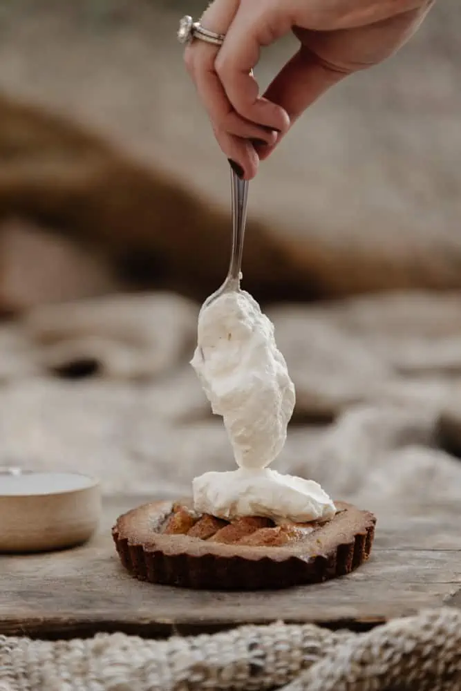 womans hand holding a spoon vertically above a pear tart with whipped cream being dolloped on top