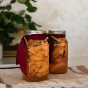 two jars of honey spiced peach preserves on a linen towel with a red ribbon tied around the top with a cinnamon stick