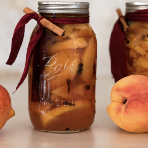 ball canning glass jar filled with peaches and spices with a red ribbon tied around the top with a cinnamon stick and whole peaches on the counter