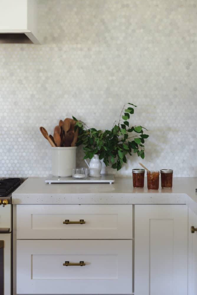 white shaker cabinets with white concrete countertops and marble penny tile backsplash kitchen with jar of wooden utensils and a plant with 3 jars of jam on the right