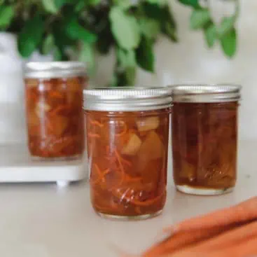 3 ball canning jars filled with carrot cake jam, the front most jar is open with a wooden spoon sticking out and plant in the background and carrots in the foreground