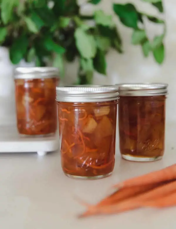3 ball canning jars filled with carrot cake jam, the front most jar is open with a wooden spoon sticking out and plant in the background and carrots in the foreground