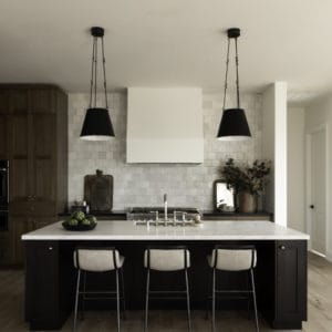 kitchen with a view looking at a dark wood island with a marble countertop and two black hanging pendents above, tiled back kitchen wall with a white hood centered.