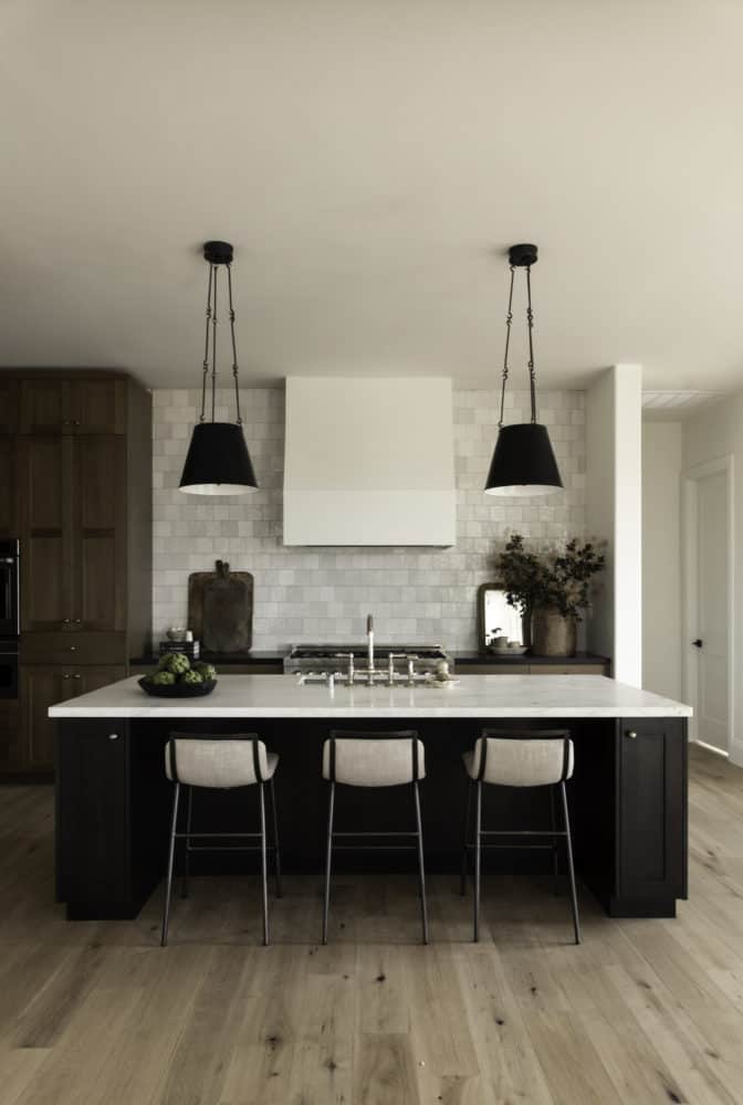 kitchen with a view looking at a dark wood island with a marble countertop and two black hanging pendents above, tiled back kitchen wall with a white hood centered.