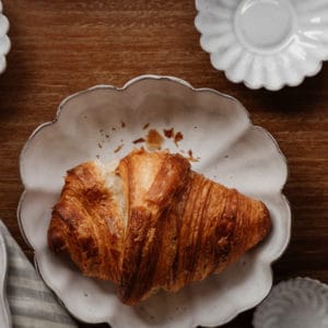 croissant on a scalloped white ceramic dish with several other white ceramic dishes surrounding it on a walnut wood surface and a striped linen towel