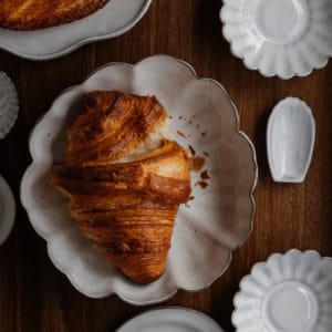 croissant on a scalloped white ceramic dish with several other white ceramic dishes surrounding it on a walnut wood surface