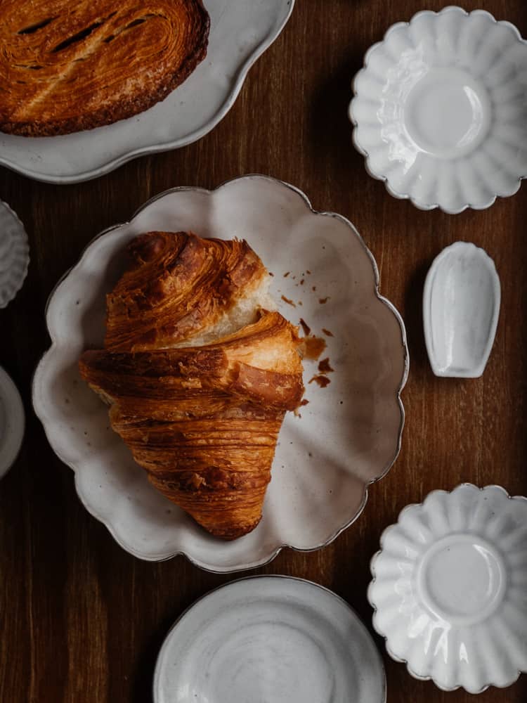 croissant on a scalloped white ceramic dish with several other white ceramic dishes surrounding it on a walnut wood surface