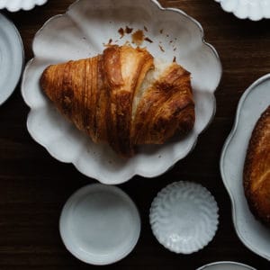 croissant on a scalloped white ceramic dish with several other white ceramic dishes surrounding it on a walnut wood surface