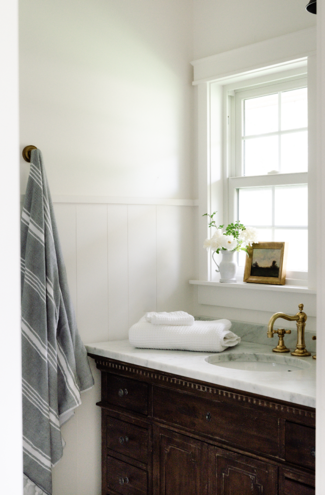 bathroom corner with a window and a dark wood vanity with marble countertops with a brass faucet and white folded towels on the counter.