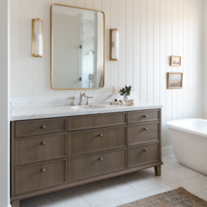 bathroom with tongue and groove walls, medium wood tone vanity with a marble countertop a brass mirror and sconces.