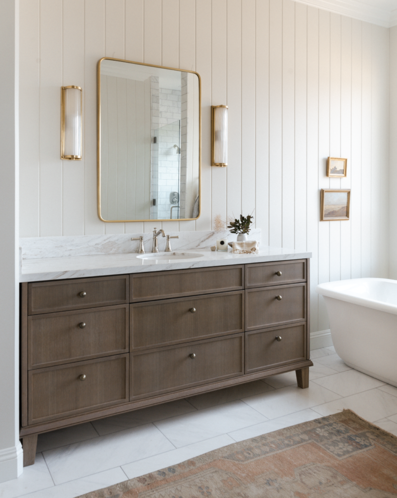 bathroom with tongue and groove walls, medium wood tone vanity with a marble countertop a brass mirror and sconces.