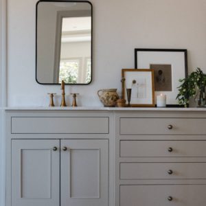 bathroom with a taupe colored vanity and marble countertops with a brass faucet, black mirror, and white and brass cone shaped sconce above. Leaning art and accessories are on the vanity countertop.