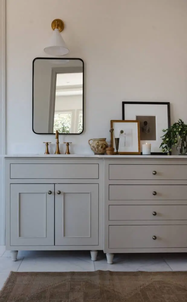 bathroom with a taupe colored vanity and marble countertops with a brass faucet, black mirror, and white and brass cone shaped sconce above. Leaning art and accessories are on the vanity countertop.