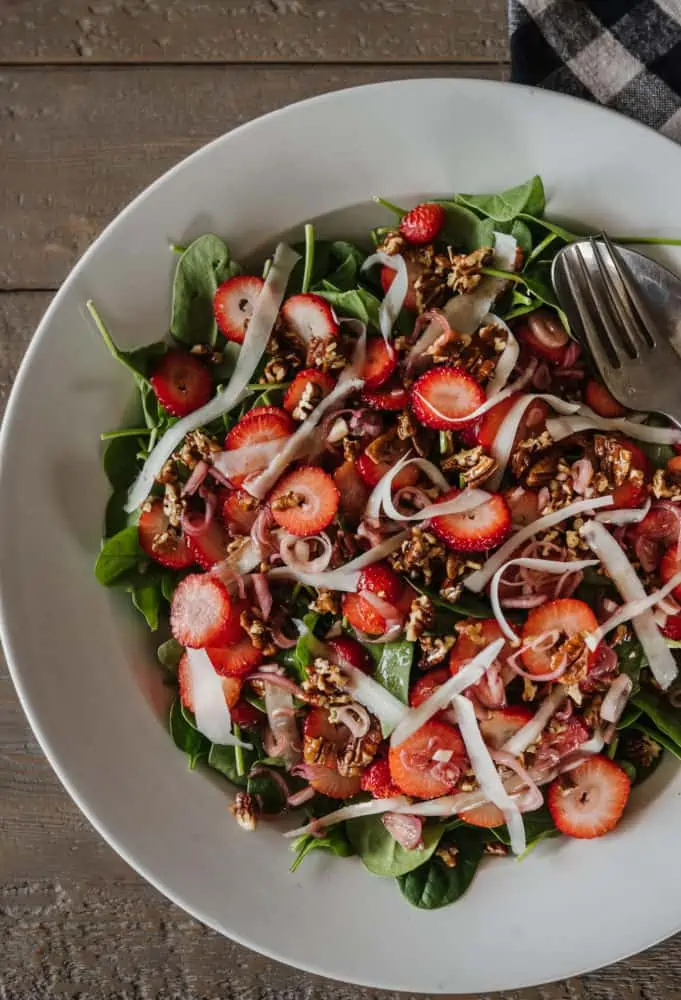 top down close up view of a large white bowl filled with strawberry spinach salad with shaved goat cheese on top and candied pecans