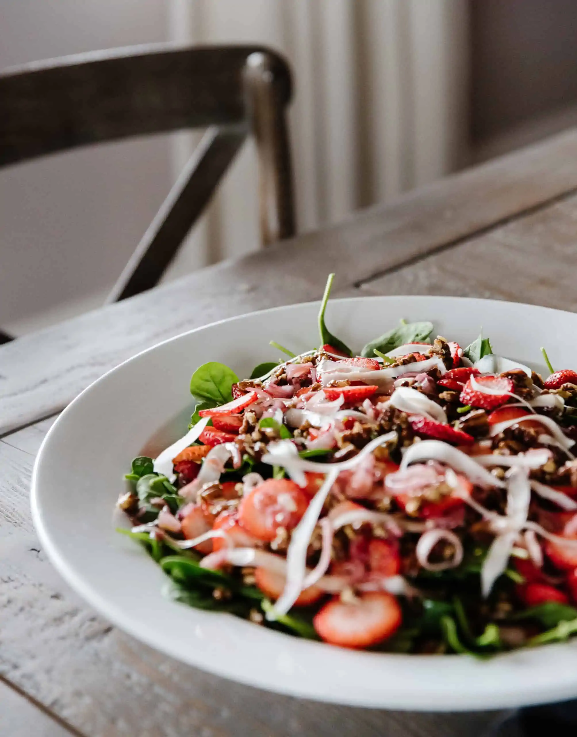 view of strawberry spinach salad in a large white bowl on a wooden table with a chair in the background