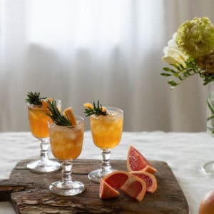 linen covered table with an antique wood cutting board with 3 clear glass goblet glasses with orange liquid inside and a rosemary sprig, cut grapefruit on the cutting board as well