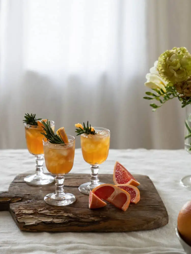 linen covered table with an antique wood cutting board with 3 clear glass goblet glasses with orange liquid inside and a rosemary sprig, cut grapefruit on the cutting board as well