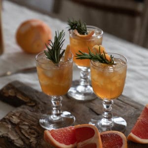 linen covered table with an antique wood cutting board with 3 clear glass goblet glasses with orange liquid inside and a rosemary sprig, cut grapefruit on the cutting board as well