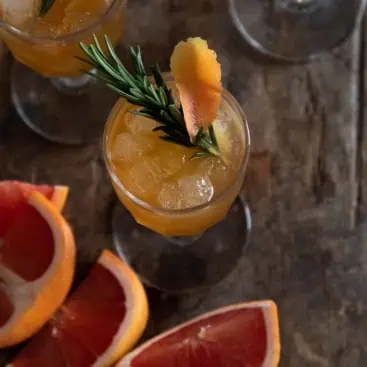 top down shot of a clear glass filled with orange liquid garnished with a rosemary sprig and cut grapefruits