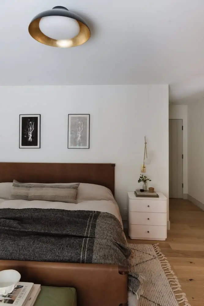 bedroom scene with a carmel eather headboard and footboard, neutral linen bedding with a charcoal throw blanket, black and white art above the headboard. White nightstand with brass and glass modern sconce next to the bed.