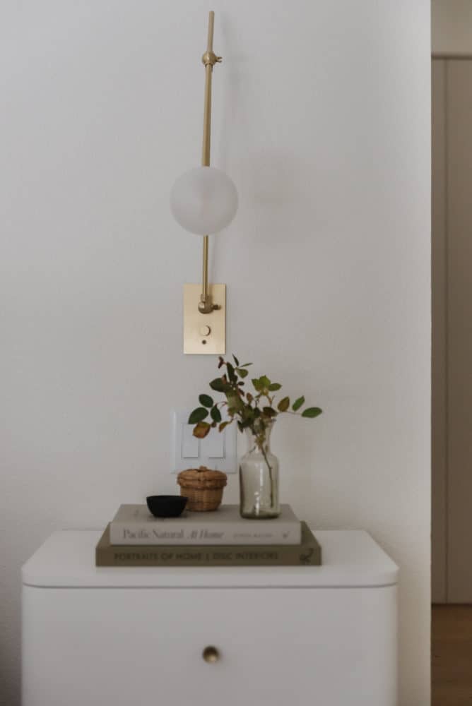 close up of white nightstand with 2 books stacked with a vase of leaves and small decorative bowls with a brass and glass modern sconce on the wall.