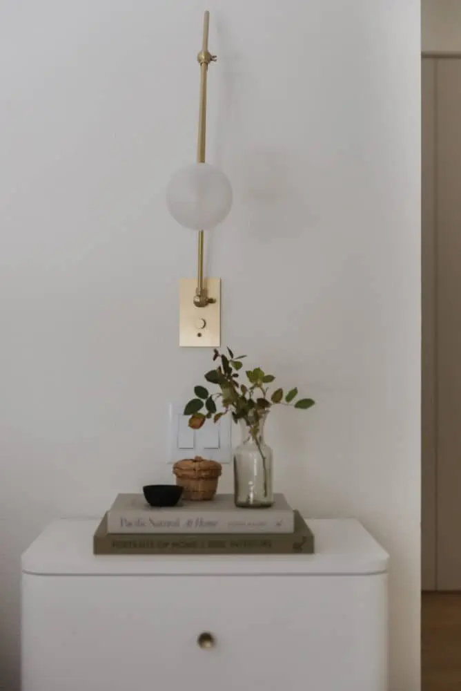 close up of white nightstand with 2 books stacked with a vase of leaves and small decorative bowls with a brass and glass modern sconce on the wall.