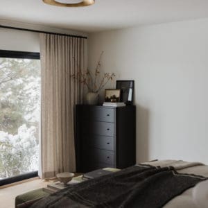 bedroom scene with medium tone wood floors, olive green velvet bench at the end of the linen duvet covered bed with a charcoal throw blanket on the bottom edge with a tall black dresser in the corner and floor to ceiling light linen curtains.