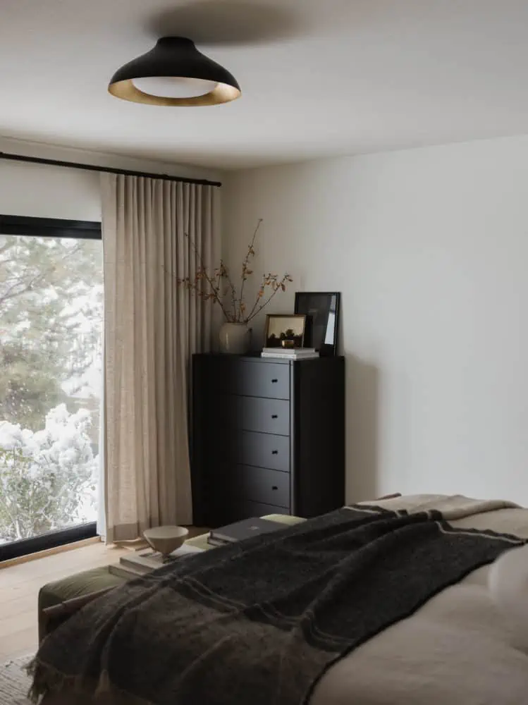 bedroom scene with medium tone wood floors, olive green velvet bench at the end of the linen duvet covered bed with a charcoal throw blanket on the bottom edge with a tall black dresser in the corner and floor to ceiling light linen curtains.