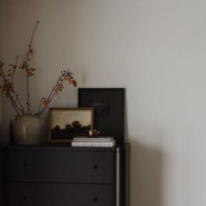 tall black dresser in a bedroom corner with linen curtain, ceramic pottery vase with tree branches inside, stacked books, and artwork.