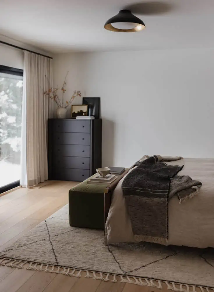 bedroom scene with medium tone wood floors, olive green velvet bench at the end of the linen duvet covered bed with a charcoal throw blanket on the bottom edge with a tall black dresser in the corner and floor to ceiling light linen curtains.