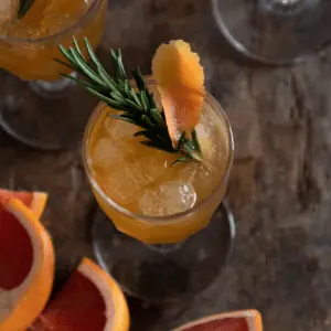 top down shot of a clear glass filled with orange liquid garnished with a rosemary sprig and cut grapefruits
