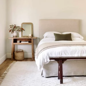 neutral bedroom scene with a light textured rug, taupe linen headboard and bedskirt, white duvet with a cream throw blanket and an olive green pillow. Light wood nightstand with plant, mirror and candle on top and a wicker basket below. Vintage wood bench at the end of the bed.