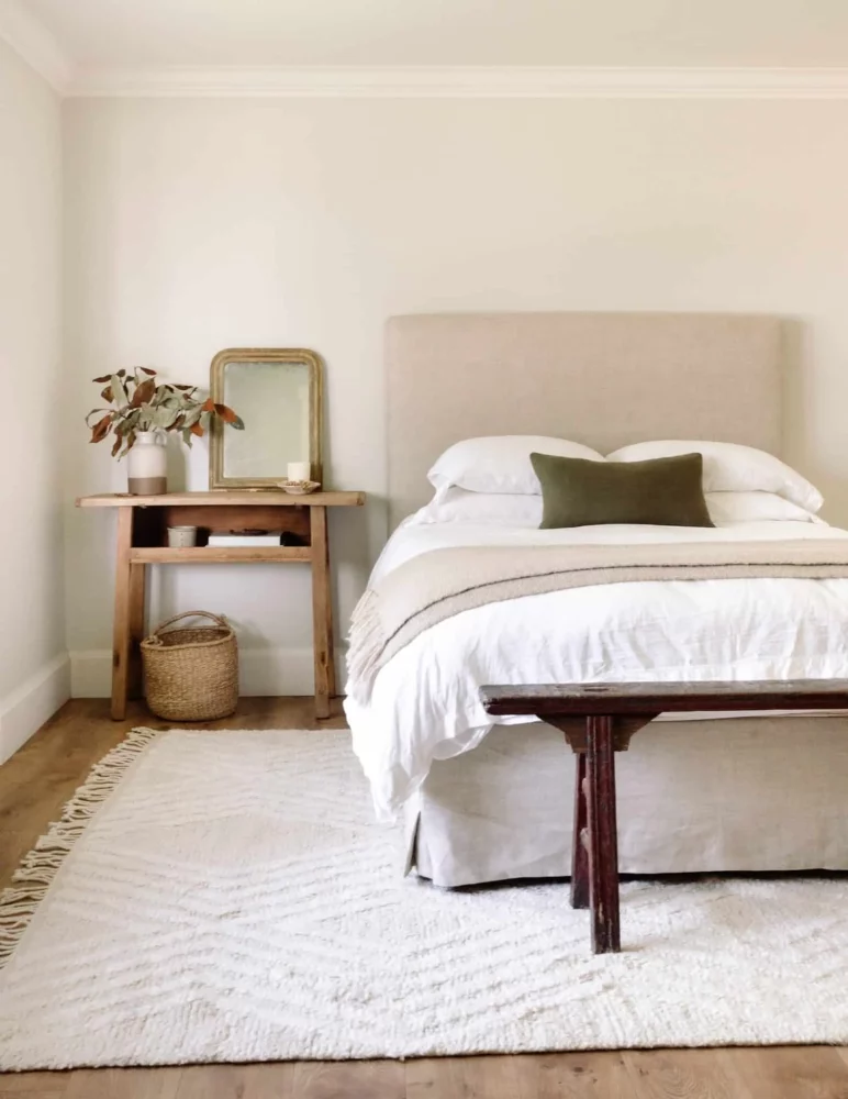 neutral bedroom scene with a light textured rug, taupe linen headboard and bedskirt, white duvet with a cream throw blanket and an olive green pillow. Light wood nightstand with plant, mirror and candle on top and a wicker basket below. Vintage wood bench at the end of the bed.